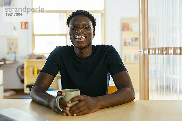 Fröhlicher junger Student  der mit einer Kaffeetasse am Schreibtisch sitzt