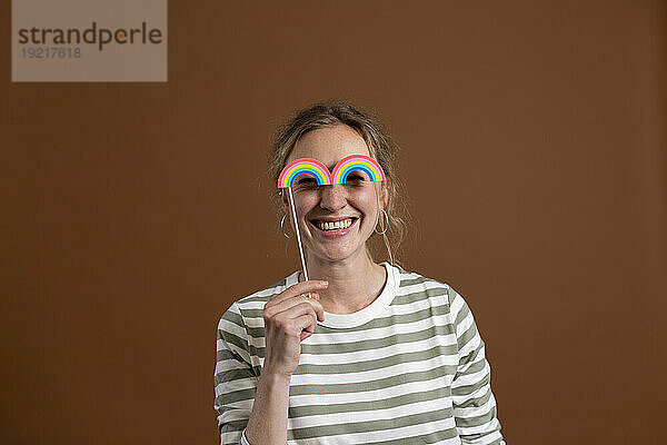 Happy woman holding rainbow prop in front of eyes against brown background