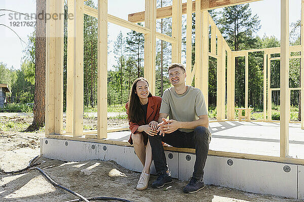 Lächelndes Paar sitzt an einem sonnigen Tag neben einem Holzkonstruktionsrahmen