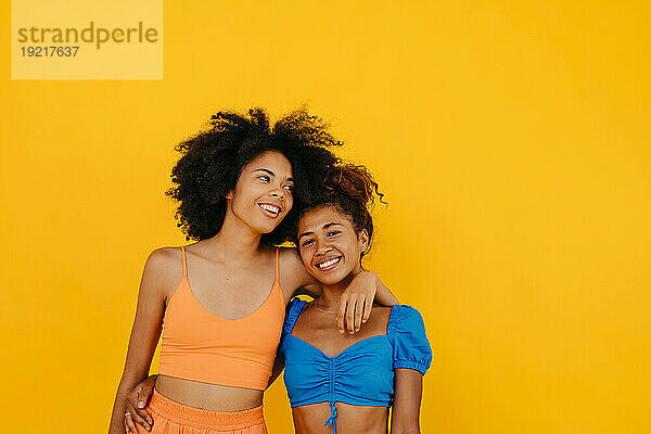 Frau mit Arm um lächelnde Freundin vor gelbem Hintergrund