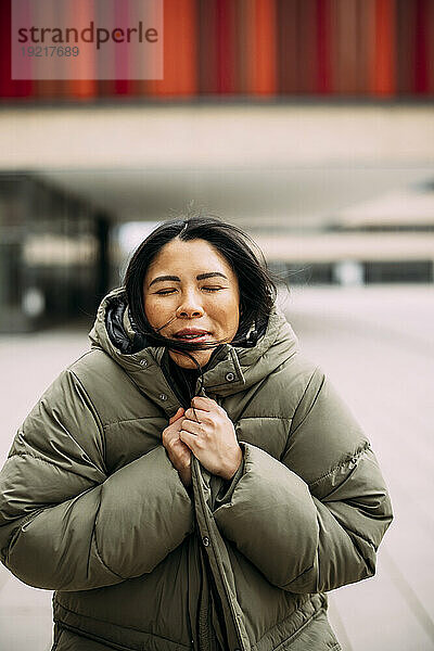 Geschäftsfrau mit geschlossenen Augen trägt gepolsterte Jacke vor Bürogebäude