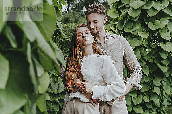 Lächelndes Paar mit geschlossenen Augen steht inmitten von Pflanzen im Garten