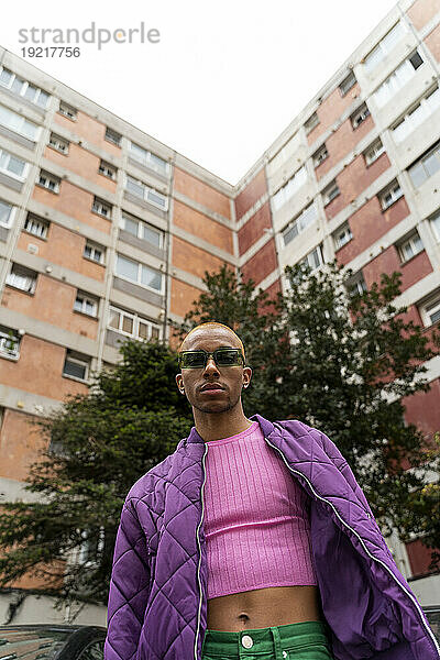 Nicht-binäre Person mit Sonnenbrille steht vor einem Gebäude