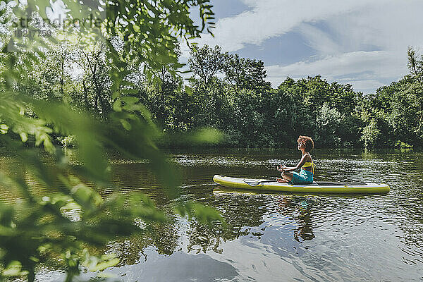 Junge Frau sitzt und meditiert auf einem Paddleboard im See