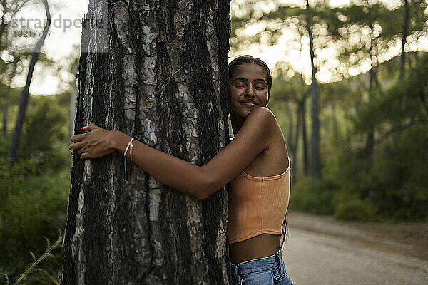 Lächelndes Teenager-Mädchen umarmt Baumstamm im Wald