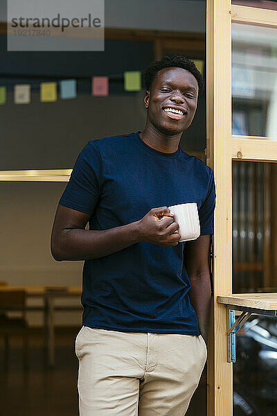 Fröhlicher junger Student steht mit Kaffeetasse im Türrahmen der Universität