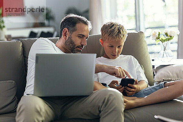 Vater und Sohn nutzen Mobiltelefon und sitzen zu Hause auf dem Sofa