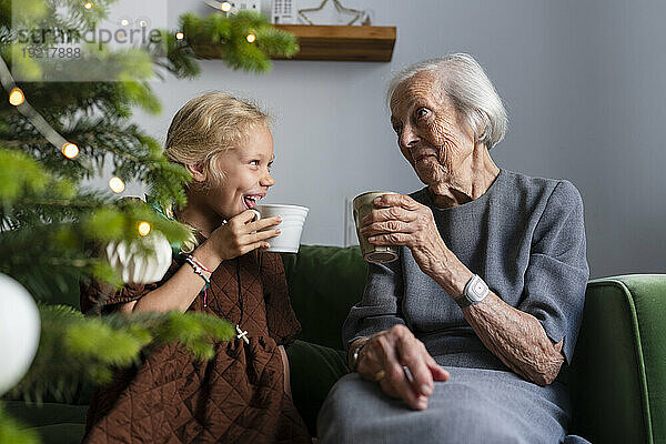Glückliches Mädchen  das mit Großmutter heiße Schokolade trinkt und seine Freizeit zu Hause in der Nähe des Weihnachtsbaums verbringt