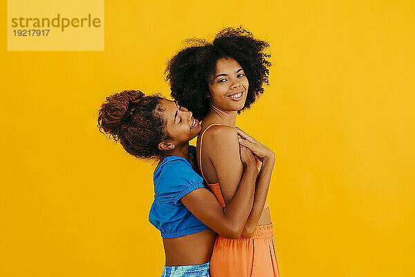 Lächelnde Frau umarmt junge Freundin vor gelbem Hintergrund