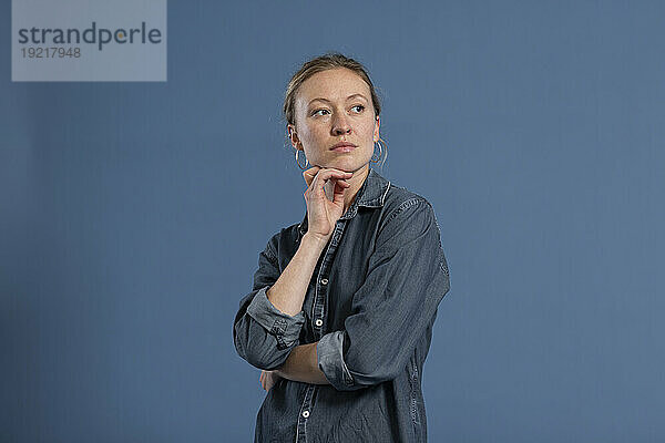 Nachdenkliche Frau mit der Hand am Kinn vor blauem Hintergrund