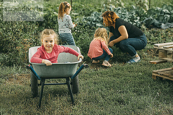 Smiling daughter sitting in wheelbarrow with mother and sisters harvesting at vegetable garden