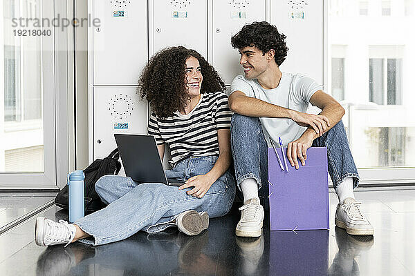 Junge Studenten sitzen mit Laptop und lachen vor dem Schließfach der Universität