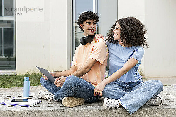 Lächelnde Studenten sitzen mit Laptop auf einer Betonbank auf dem Campus