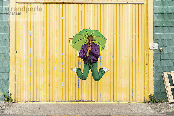 Nicht-binäre Person springt mit grünem Regenschirm vor gelber Fensterladentür