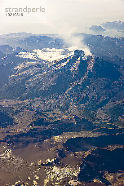 Luftaufnahme des Mount Redoubt mit Dampf  der aus dem Krater kommt  Vulkanasche auf dem Eisfeld im Vordergrund und Mt. Iliamna in weiter Ferne  Südwest-Alaska