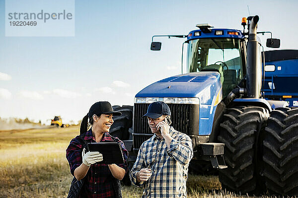 Ein Mann und eine Frau verwalten und überwachen ihre Rapsernte mit tragbaren drahtlosen Geräten und tätigen einen Anruf  während sie vor einem großen Traktor stehen. Alcomdale  Alberta  Kanada