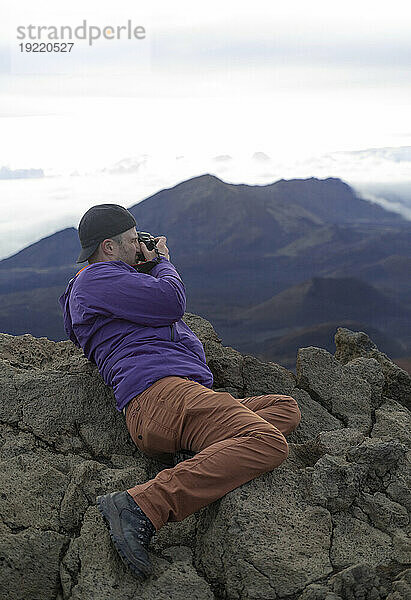 Mann positioniert sich auf den Vulkangesteinen auf einem Berggipfel über den Wolken bei Haleakala  beobachtet den Sonnenaufgang über der Pazifikküste und fotografiert die Aussicht; Haleakala-Nationalpark  Maui  Hawaii  Vereinigte Staaten von Amerika