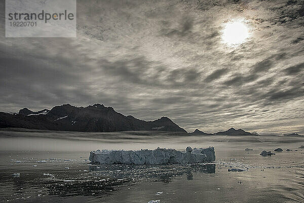 Ein im Gletscherwasser des Nansen-Fjords schwimmender Eisberg mit Silhouetten der Berge vor der Sonne  die durch die grauen Wolken scheint  und einer Nebelschicht über dem ruhigen Wasser; Ostgrönland  Grönland
