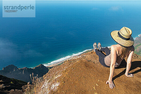 Blick von hinten auf eine Frau mit Strohhut  die auf der Spitze einer Bergklippe sitzt und auf das strahlend blaue Wasser des Pazifischen Ozeans blickt  auf dem Kalalau Trail entlang der Napali-Küste; Kauai  Hawaii  Vereinigte Staaten von Amerika