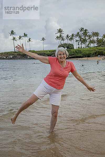 Porträt einer reifen Frau  die in einer verspielten Position im Wasser steht und am Sandstrand des Kapalua Resorts in West Maui in die Kamera lächelt; Maui  Hawaii  Vereinigte Staaten von Amerika