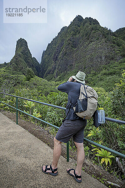 Blick von hinten auf einen Mann  der auf einem Gehweg steht und die Aussicht auf den Kuka‘emoku  Iao Needle im üppigen Iao-Tal im Zentrum von Maui fotografiert; Maui  Hawaii  Vereinigte Staaten von Amerika
