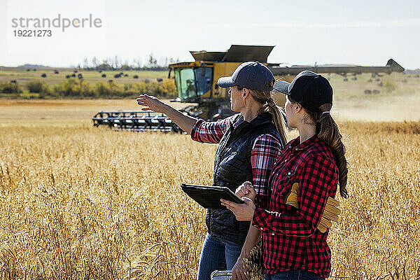 Eine reife Bäuerin steht auf einem Feld und arbeitet zur Erntezeit mit einer jungen Frau zusammen  nutzt fortschrittliche Agrarsoftware auf einem Pad und beobachtet dabei einen Mähdrescher bei der Arbeit im Hintergrund. Alcomdale  Alberta  Kanada