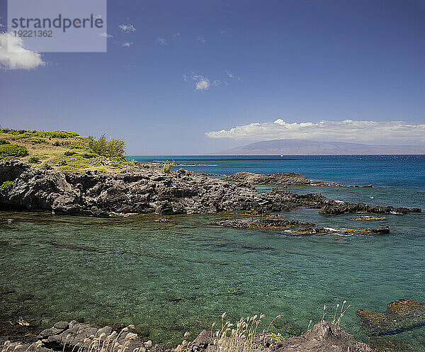 Nahaufnahme des klaren  türkisfarbenen Wassers entlang der felsigen Küste mit Fernblick auf ein am Horizont festgemachtes Boot und die Silhouetten von Bergen unter einem blauen Himmel; Maui  Hawaii  Vereinigte Staaten von Amerika