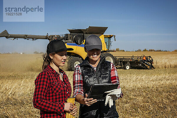 Eine reife Bäuerin steht auf einem Feld und arbeitet gemeinsam mit einer jungen Frau bei der Ernte mithilfe fortschrittlicher landwirtschaftlicher Software auf einem Pad  während im Hintergrund ein Mähdrescher arbeitet. Alcomdale  Alberta  Kanada