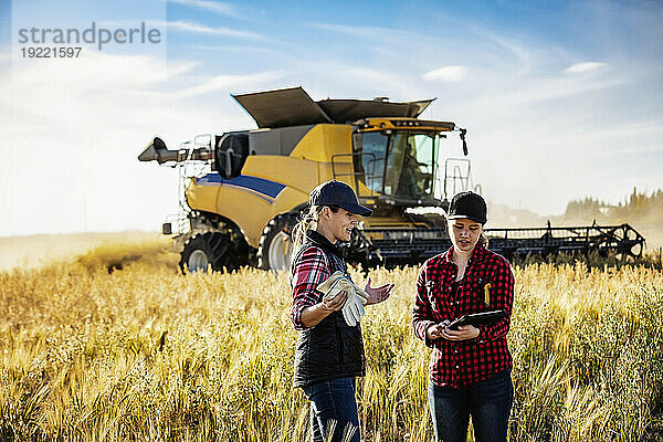 Eine reife Bäuerin  die auf einem Feld steht und mit einer jungen Frau zur Erntezeit zusammenarbeitet  wobei sie fortschrittliche landwirtschaftliche Software auf einem Pad verwendet  während im Hintergrund ein Mähdrescher arbeitet; Alcomdale  Alberta  Kanada