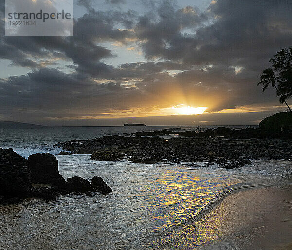 Sonnenstrahlen strahlen durch die Wolken und spiegeln sich im Wasser am Secret Beach mit Blick auf die Bucht und den Pazifischen Ozean in der Dämmerung mit Silhouetten von Menschen  die am felsigen Ufer entlang spazieren; Makena  Maui  Hawaii  Vereinigte Staaten von Amerika