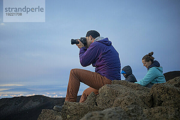 Familie sitzt auf einem Berggipfel über den Wolken bei Haleakala und beobachtet den Sonnenaufgang über der Pazifikküste  während der Vater die Aussicht fotografiert; Haleakala-Nationalpark  Maui  Hawaii  Vereinigte Staaten von Amerika