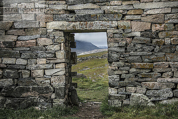 Nahaufnahme einer Steinmauer mit einer Öffnung  die einen malerischen Ausblick auf die archäologische Stätte Hvalsey in der Nähe von Qaqortoq bietet  wo sich an der Südspitze Grönlands im Nordatlantik eine antike Ruine einer Kirche aus dem Jahr 1300 n. Chr. befindet; Südgrönland  Grönland