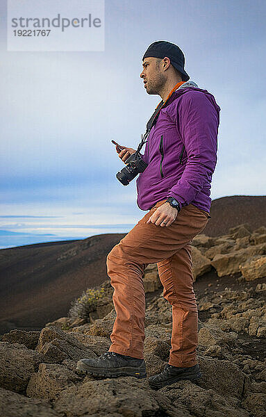 Mann  der auf Vulkangestein auf einem Berggipfel über den Wolken bei Haleakala steht und den Sonnenaufgang über der Pazifikküste beobachtet  ein Smartphone mit einer Kamera um den Hals hält und die Aussicht fotografiert; Haleakala-Nationalpark  Maui  Hawaii  Vereinigte Staaten von Amerika