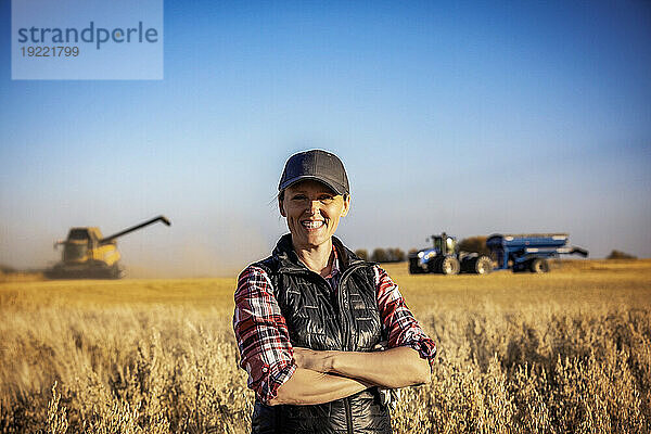 Porträt einer reifen Bäuerin  die auf einem Getreidefeld steht  während der Ernte für die Kamera posiert und lächelt  während im Hintergrund bei Sonnenuntergang ein Mähdrescher sich darauf vorbereitet  eine gemischte Ernte auf einen Getreidewagen abzuladen; Alcomdale  Alberta  Kanada