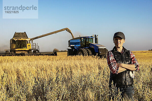 Porträt einer reifen Bäuerin  die auf einem Getreidefeld steht und während der Ernte für die Kamera posiert  während im Hintergrund bei Sonnenuntergang ein Mähdrescher eine gemischte Ernte auf einen Getreidewagen entlädt; Alcomdale  Alberta  Kanada