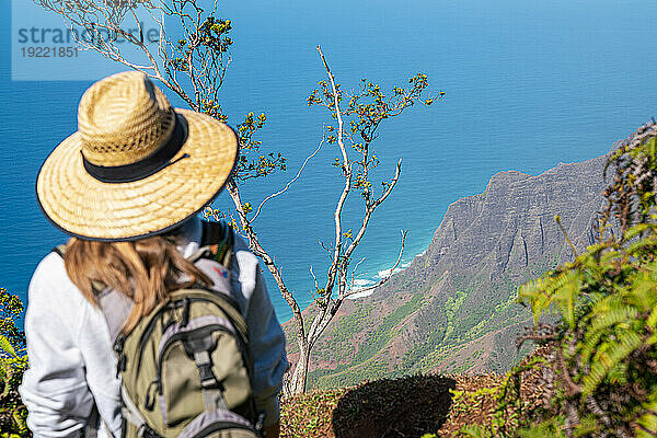 Blick von hinten auf eine Frau mit Strohhut  die auf der Spitze einer Bergklippe steht und auf das strahlend blaue Wasser des Pazifischen Ozeans blickt  auf dem Kalalau Trail entlang der Napali-Küste; Kauai  Hawaii  Vereinigte Staaten von Amerika