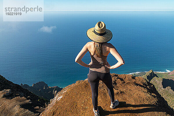 Blick von hinten auf eine Frau mit Strohhut  die auf der Spitze einer Bergklippe steht und auf das strahlend blaue Wasser des Pazifischen Ozeans entlang des Kalalau Trail an der Napali-Küste blickt; Kauai  Hawaii  Vereinigte Staaten von Amerika