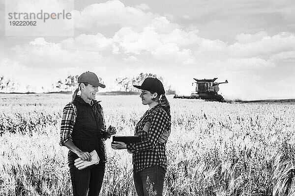 Eine reife Bäuerin  die auf einem Feld steht und mit einer jungen Frau zur Erntezeit zusammenarbeitet  wobei sie fortschrittliche landwirtschaftliche Software auf einem Pad verwendet  während im Hintergrund ein Mähdrescher arbeitet; Alcomdale  Alberta  Kanada