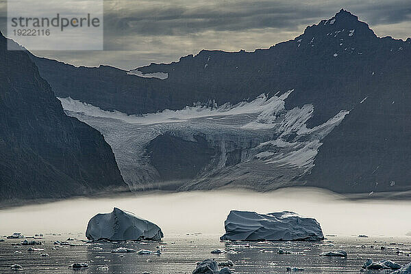 Im Nebel schwebende Eisberge vor dem Gletscher am Nansen-Fjord mit der Silhouette der schroffen Berggipfel vor einem grauen  bewölkten Himmel im Hintergrund; Ostgrönland  Grönland