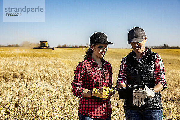 Eine reife Bäuerin steht auf einem Feld und arbeitet gemeinsam mit einer jungen Frau bei der Ernte mithilfe fortschrittlicher landwirtschaftlicher Software auf einem Pad  während im Hintergrund ein Mähdrescher arbeitet. Alcomdale  Alberta  Kanada