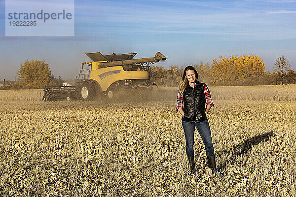 Porträt einer reifen Bäuerin  die auf einem Getreidefeld steht und während der Ernte für die Kamera posiert  während im Hintergrund bei Sonnenuntergang ein Mähdrescher arbeitet; Alcomdale  Alberta  Kanada