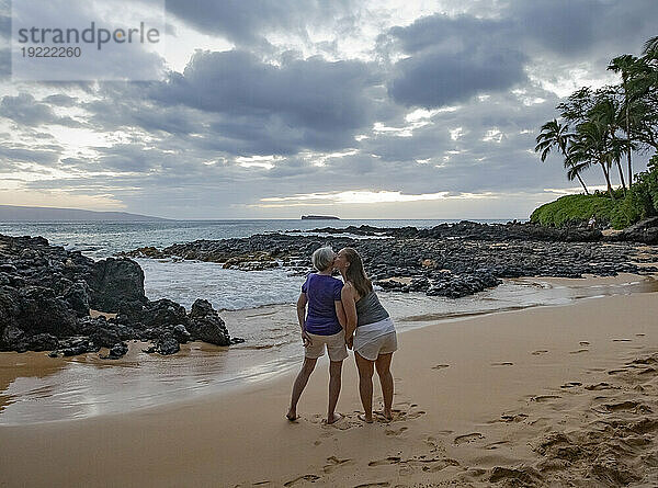 Blick von hinten auf eine reife Mutter mit ihrer erwachsenen Tochter  die Händchen haltend auf dem Sand des Secret Beach steht und in der Dämmerung auf die Bucht und den Pazifischen Ozean blickt; Makena  Maui  Hawaii  Vereinigte Staaten von Amerika