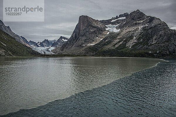 Blick auf den Gletscher  der durch die Berggipfel an der Südspitze Grönlands fließt  mit zwei Wasserarten  die sich am Gletscherfuß im Prins Christian Sund unter einem grauen Himmel vermischen; Südgrönland  Grönland