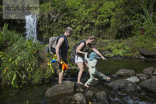 Eine Familie geht vorsichtig auf einem Pfad aus Vulkangestein  während sie auf der Straße nach Hana einen Bach mit einem wunderschönen Wasserfall im Hintergrund überquert; Maui  Hawaii  Vereinigte Staaten von Amerika