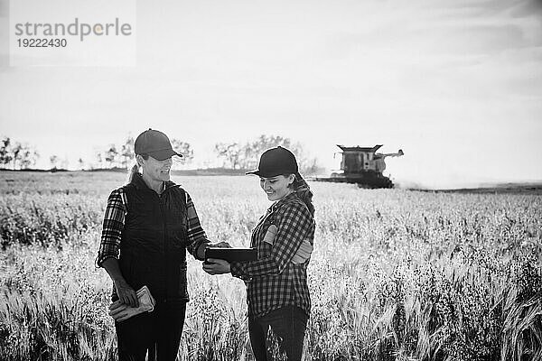 Eine reife Bäuerin  die auf einem Feld steht und mit einer jungen Frau zur Erntezeit zusammenarbeitet  wobei sie fortschrittliche landwirtschaftliche Software auf einem Pad verwendet  während im Hintergrund ein Mähdrescher arbeitet; Alcomdale  Alberta  Kanada