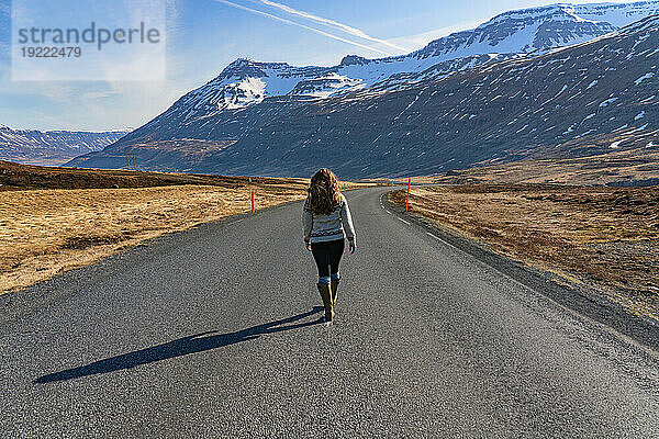 Frau geht eine offene Straße entlang in Richtung Seyðisfjörður mit Blick auf die schneebedeckten Berge und die schroffe Schönheit der Ostfjorde unter blauem Himmel; Ostisland  Island