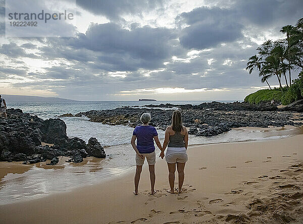 Blick von hinten auf eine reife Mutter mit ihrer erwachsenen Tochter  die Händchen haltend auf dem Sand des Secret Beach steht und in der Dämmerung auf die Bucht und den Pazifischen Ozean blickt; Makena  Maui  Hawaii  Vereinigte Staaten von Amerika