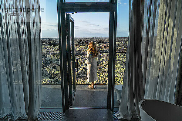 Frau steht im Bademantel auf dem Balkon und blickt auf die Lavafelder im Süden Islands; Blaue Lagune  Südisland  Island