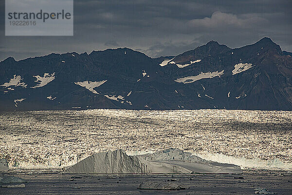 Spätlicht auf dem Gletscher am Nansen-Fjord mit der Silhouette der zerklüfteten Berggipfel im Hintergrund unter einem grauen  wolkigen Himmel; Ostgrönland  Grönland