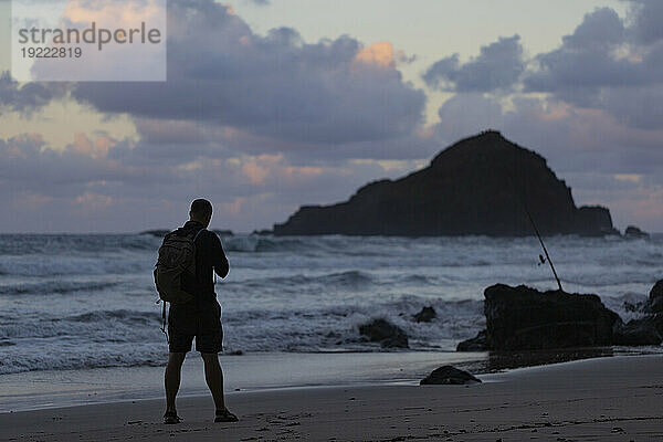 Blick von hinten auf einen silhouettierten Mann  der in der Dämmerung auf der Straße nach Hana am Strand steht und auf den Pazifischen Ozean blickt; Maui  Hawaii  Vereinigte Staaten von Amerika